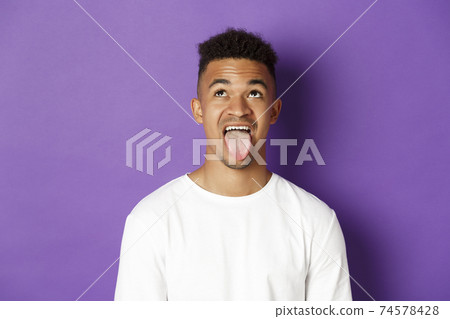 Close-up of silly african-american guy showing tongue, looking up at something lame, standing over purple background Close-up of silly african-american guy showing tongue, looking up at something lame, standing over purple background 74578428