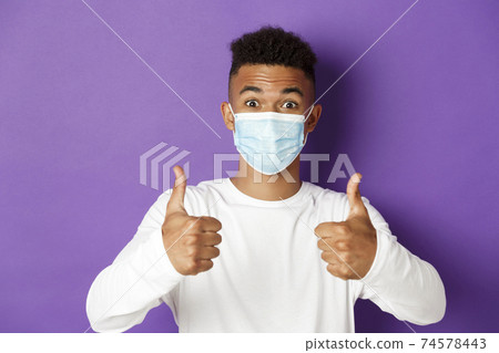 Concept of coronavirus, quarantine and social distancing. Close-up of young african-american guy in medical mask, looking excited and recommending something, showing thumbs-up in approval 74578443
