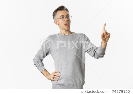 Image of confident middle-aged businessman in grey sweater and glasses, pointing finger and making speech, giving tasks to employees, standing over white background Image of confident middle-aged businessman in grey sweater and glasses, pointing finger and making speech, giving tasks to employees, standing over white background 74579100