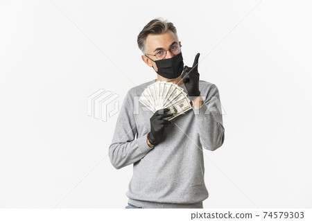Concept of covid-19, social distancing and lifestyle. Portrait of lucky middle-aged man in medical mask, gloves and glasses, showing money and pointing finger up, smiling pleased 74579303