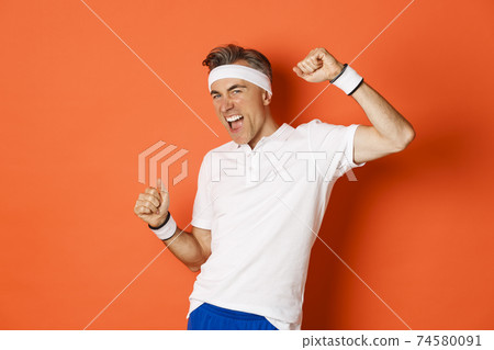 Portrait of cheerful middle-aged male athlete, become champion, raising hands up and shouting for joy, doing sports over orange background Portrait of cheerful middle-aged male athlete, become champion, raising hands up and shouting for joy, doing sports over orange background 74580091