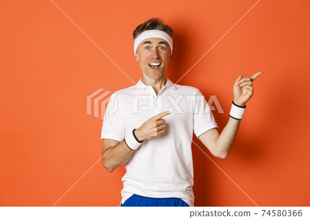 Concept of workout, sports and lifestyle. Portrait of handsome middle-aged man in sportswear, pointing fingers at upper right corner and looking amazed, showing logo, orange background 74580366