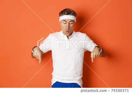Concept of workout, sports and lifestyle. Amazed and excited middle-aged sportsman in fitness clothing, pointing fingers down and looking amused at logo, standing over orange background 74580371