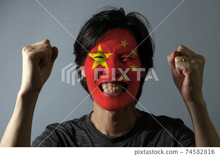 Cheerful portrait of a man with the flag of the China painted on his face on grey background. Cheerful portrait of a man with the flag of the China painted on his face on grey background. 74582816