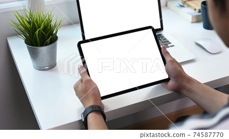 Cropped shot of young man holding digital tablet with white screen and using computer laptop at home office room. 74587770