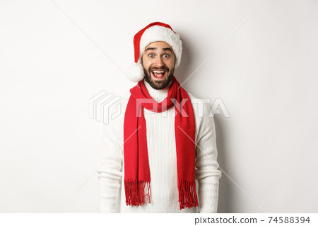 Christmas holidays. Bearded man looking surprised at camera, wearing santa party hat and red scarf, standing against white background 74588394