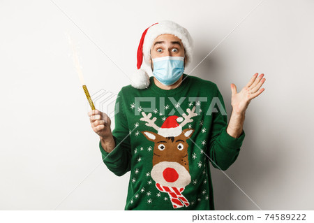 Concept of covid-19 and Christmas holidays. Happy young man in face mask celebrating Christmas, holding party sparkler and rejoicing, standing over white background 74589222