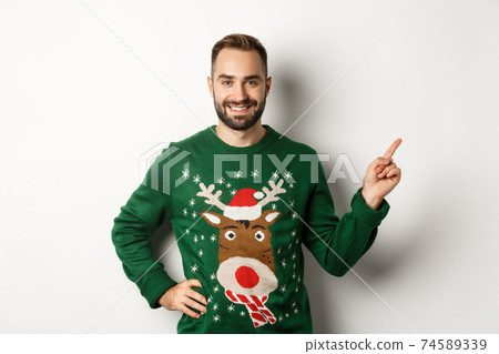 Winter holidays and christmas. Handsome bearded adult man pointing finger right at promo banner, standing in funny sweater against white background Winter holidays and christmas. Handsome bearded adult man pointing finger right at promo banner, standing in funny sweater against white background 74589339