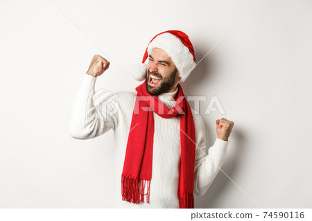 Winter holidays and New Year party concept. Handsome bearded man in santa hat winning prize, achieve goal and celebrating, making fist pump and saying yes, white background 74590116