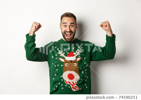 New Year, holidays and celebration. Relieved and excited man in Christmas sweater, raising hands up and celebrating, triumphing, standing over white background New Year, holidays and celebration. Relieved and excited man in Christmas sweater, raising hands up and celebrating, triumphing, standing over white background 74590251