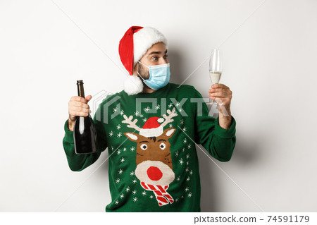 Concept of covid-19 and Christmas holidays. Man in medical mask and Santa hat enjoying glass of champagne, celebrating New Year, standing over white background 74591179