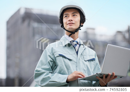 一個穿著工作服的人,看著在建築工地前的一台計算機 一個穿著工作服的人,看著在建築工地前的一台計算機 74592805