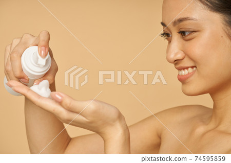 Close up portrait of pretty young woman smiling, holding a bottle of gentle foam facial cleanser isolated over beige background Close up portrait of pretty young woman smiling, holding a bottle of gentle foam facial cleanser isolated over beige background 74595859