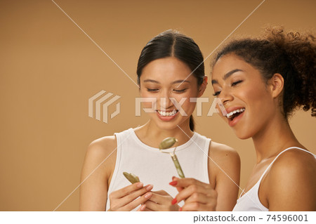 Smiling young women holding, looking at jade roller and facial gua sha while posing together isolated over beige background Smiling young women holding, looking at jade roller and facial gua sha while posing together isolated over beige background 74596001
