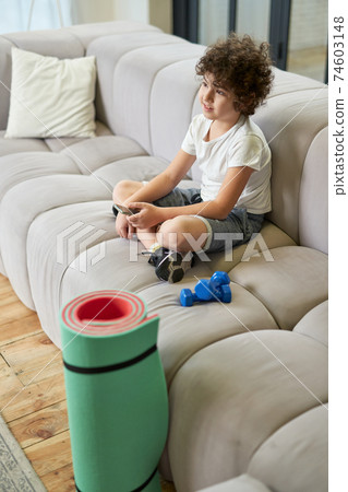 Cute latin boy sitting on a sofa and using smartphone instead of doing sports while spending time at home Cute latin boy sitting on a sofa and using smartphone instead of doing sports while spending time at home 74603148
