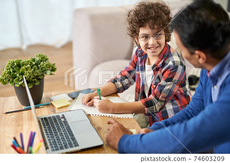 Cute learner. Portrait of joyful little hispanic school boy doing homework together with his father, using laptop while sitting at the desk at home 74603209