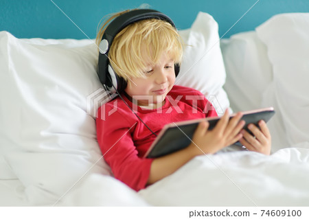 Cute preschool boy wearing headphones watches cartoon movie by tablet in bed Cute preschool boy wearing headphones watches cartoon movie by tablet in bed 74609100