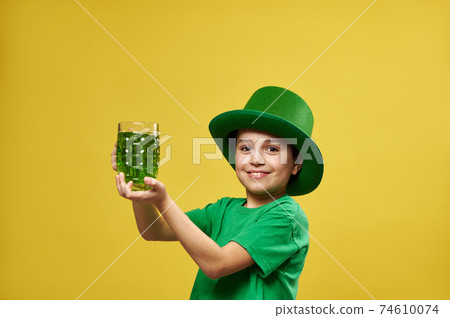 Funny little boy wearing leprechaun Irish green hat holds a glass with green drink and smiles on camera. Saint Patrick's Day 74610074