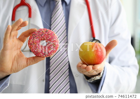 Doctor in office holding in hand pink doughnut vs red apple Doctor in office holding in hand pink doughnut vs red apple 74611472
