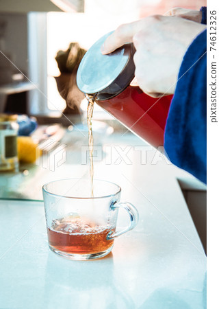 Close up man in home bathrobe pouring hot tea from glass teapot into cup on the kitchen table. Process brewing tea in the morning. A cup of freshly brewed black tea, warm soft light. Selective focus. Close up man in home bathrobe pouring hot tea from glass teapot into cup on the kitchen table. Process brewing tea in the morning. A cup of freshly brewed black tea, warm soft light. Selective focus. 74612326