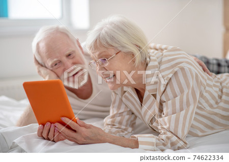 Grey-haired woman showing something on a tablet to her husband Grey-haired woman showing something on a tablet to her husband 74622334