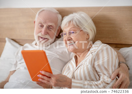 Aged married couple lying in bed and having good time while reading Aged married couple lying in bed and having good time while reading 74622336