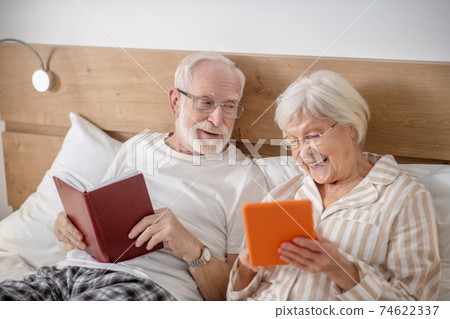 Aged married couple lying in bed and having good time while reading Aged married couple lying in bed and having good time while reading 74622337