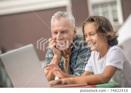 Son and dad sitting together with a laptop and looking contented Son and dad sitting together with a laptop and looking contented 74622418