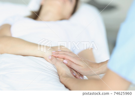 On bed lies a sick doctor sitting next to him and sympathetically holds hand 74625960