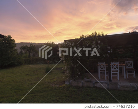 View of summer garden with tiber wooden gazebo pergola with blooming climbing plant flowers and white furniture and surrounding sandstone wall, green grass and bush background and vivid sunset sky. 74627210