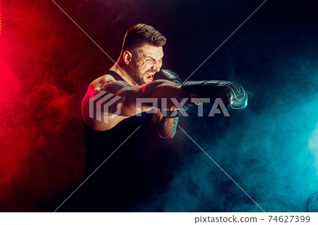 Bearded tattooed sportsman muay thai boxer in black undershirt and boxing gloves fighting on dark background with smoke. Bearded tattooed sportsman muay thai boxer in black undershirt and boxing gloves fighting on dark background with smoke. 74627399