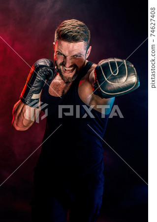 Bearded tattooed sportsman muay thai boxer in black undershirt and boxing gloves fighting on dark background with smoke. 74627408