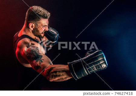 Bearded tattooed sportsman muay thai boxer in black undershirt and boxing gloves fighting on dark background with smoke. 74627418