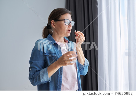 Mature middle-aged woman at home holding pill and glass of fresh water Mature middle-aged woman at home holding pill and glass of fresh water 74630992