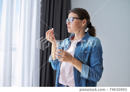 Mature middle-aged woman at home holding pill and glass of fresh water Mature middle-aged woman at home holding pill and glass of fresh water 74630993