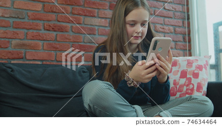 Little girl preschool child using a digital smartphone technology device looks telephone. Plays in the phone sitting on the sofa alone. Little girl preschool child using a digital smartphone technology device looks telephone. Plays in the phone sitting on the sofa alone. 74634464