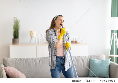 House cleaning is fun. Cheerful young lady using mop as mic, singing song while doing cleanup at living room 74634604