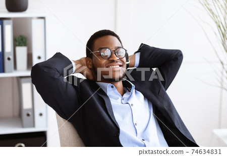 Joyful black businessman reclining on chair in his cabinet 74634831