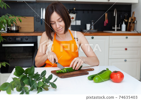 Young beautiful woman in apron sitting at table, cooking at home, chopping vegetables by knife 74635403