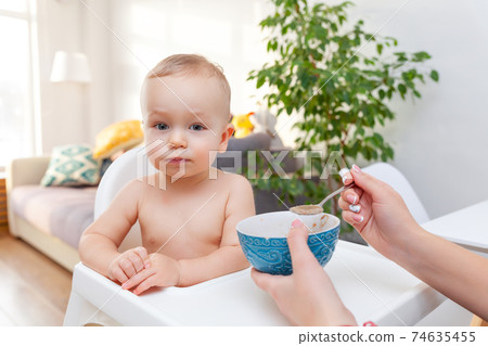 Mother's hand feeds funny cute happy blond baby toddler with spoon 74635455