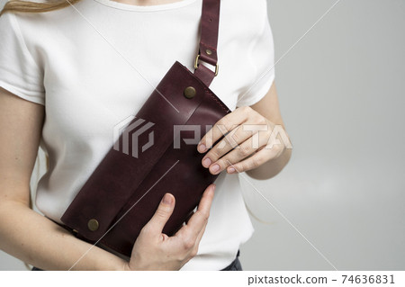 Girl in a white blouse with a leather red handmade bag over her shoulder. Designer dark red banana bag. Woman in a studio. Comfortable small bag for walking. 74636831