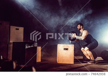 Fit tattoed bearded man jumping onto a box as part of exercise routine. Man doing box jump in the gym. Athlete is performing box jumps 74642938