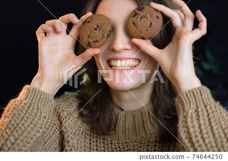 Young girl in knitted beige sweater is holding two chocolate cookies covering her eyes on black background. 74644250