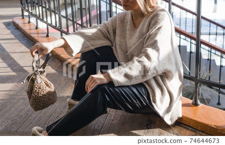 A stylish girl in black trousers and a light sweater sits on the floor and holds a bag in the room against the background of large windows. Close-up. A stylish girl in black trousers and a light sweater sits on the floor and holds a bag in the room against the background of large windows. Close-up. 74644673