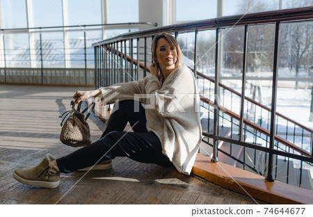 A stylish girl in black trousers and a light sweater sits on the floor and holds a bag in the room against the background of large windows. Close-up. A stylish girl in black trousers and a light sweater sits on the floor and holds a bag in the room against the background of large windows. Close-up. 74644677