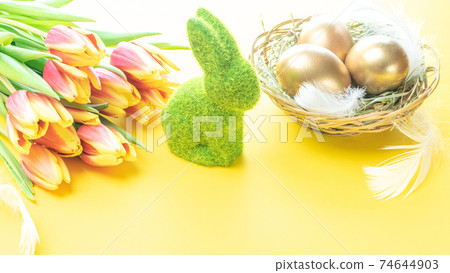 Easter eggs gold. Golden colour egg in basket with spring tulips, white feathers on pastel yellow background in Happy Easter decoration. Congratulatory easter design. Easter eggs gold. Golden colour egg in basket with spring tulips, white feathers on pastel yellow background in Happy Easter decoration. Congratulatory easter design. 74644903