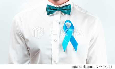 Blue ribbon cancer. Awareness prostate of health in November. Hipster men in bright shirt, cyan bowtie with blue ribbon in hands on white background. Symbol of oncology affected man. Copy space. Blue ribbon cancer. Awareness prostate of health in November. Hipster men in bright shirt, cyan bowtie with blue ribbon in hands on white background. Symbol of oncology affected man. Copy space. 74645041