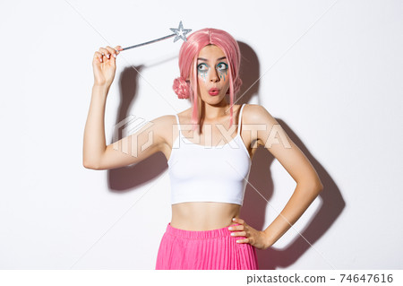 Image of silly attractive girl in pink wig, dressing as fairy for halloween party, hoding magic wand, standing over white background 74647616