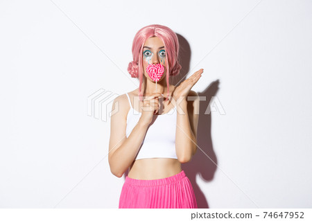 Image of flirty female model with pink wig and bright makeup, kissing heart-shaped candy and looking surprised, celebrating halloween 74647952
