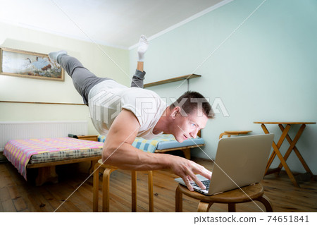 A muscular man practice yoga on one hand and works at a laptop. Self-isolation, remote working and healthy lifestyle concept A muscular man practice yoga on one hand and works at a laptop. Self-isolation, remote working and healthy lifestyle concept 74651841